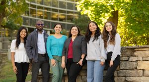 Six people stand in front of stone wall on UW Madison campus
