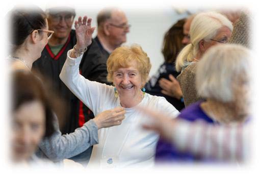 The image depicts an indoor social gathering or event. At the center, a person in a white shirt is raising one arm while another individual appears to be adjusting something on their shirt. Several other people are present in the background, engaged in conversation or other activities.