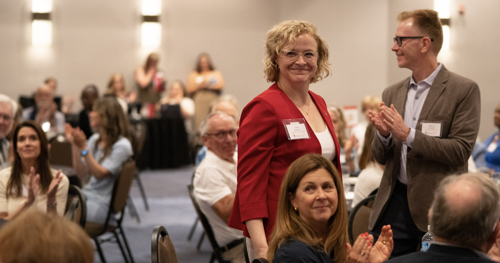 Woman in red blazer and man in gray suit stand in ballroom with audience clapping