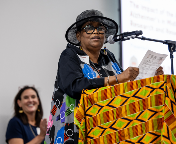 Person stands at a podium wearing a black hat and speaks to a smiling audience