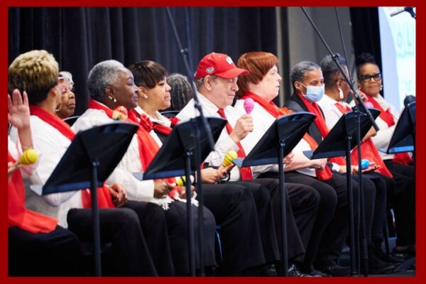 A group of chorus members sit in chairs with microphones and music stands on a stage
